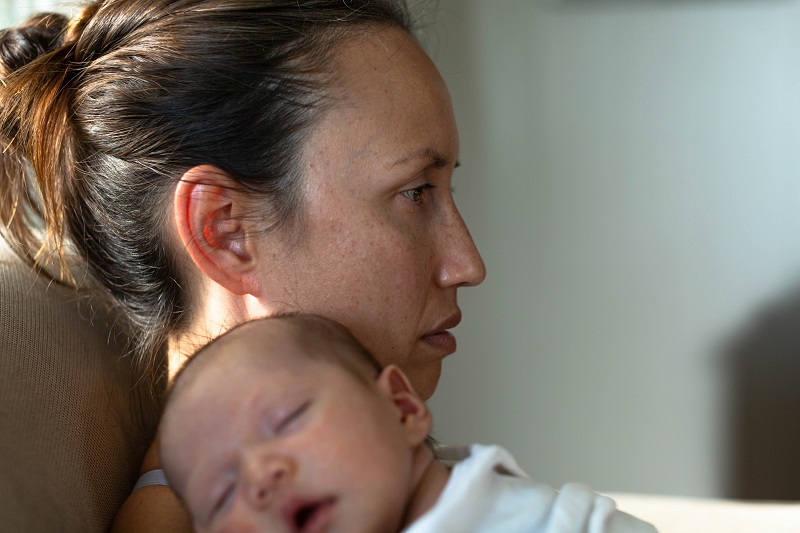 A mom experiencing the "baby blues", feeling unhappy, holding her baby at home sitting on the couch thinking.