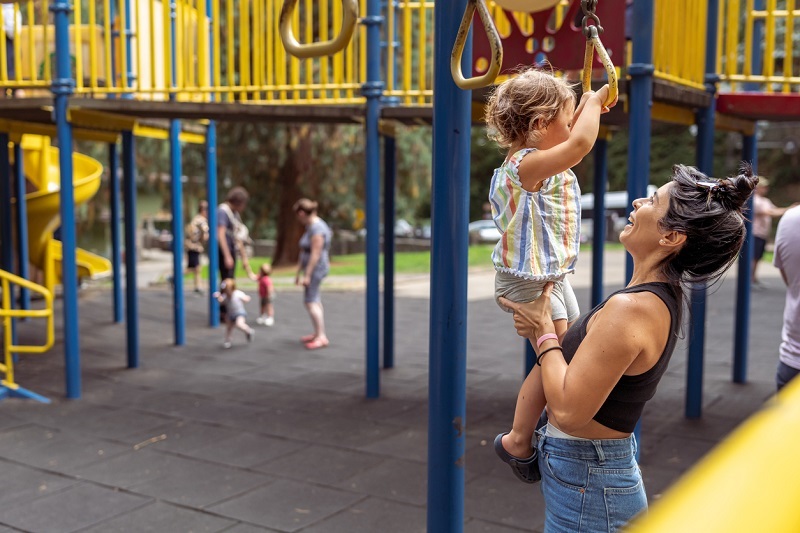 Mom helps a two year old girl use the jungle gym at the playground on a warm sunny afternoon.