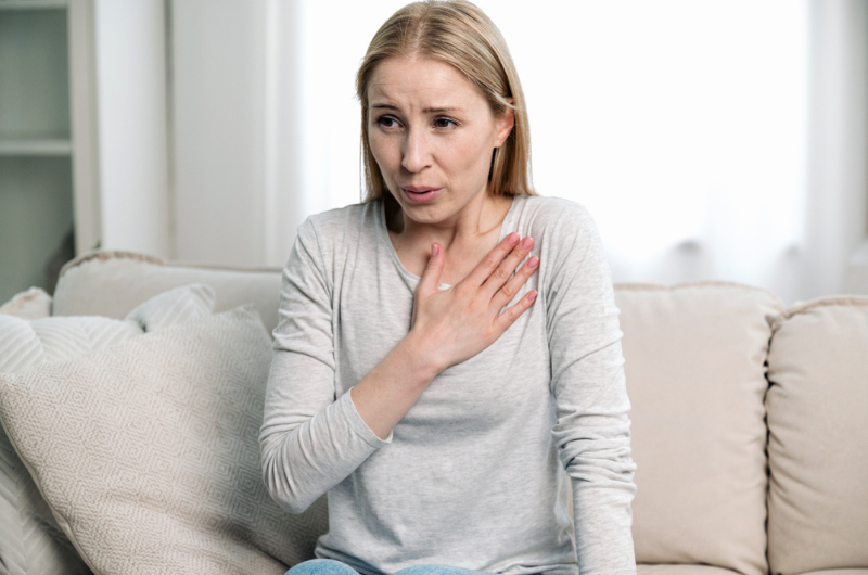 Young woman holds her hand on her chest and has difficulty breathing while sitting on a couch at home.