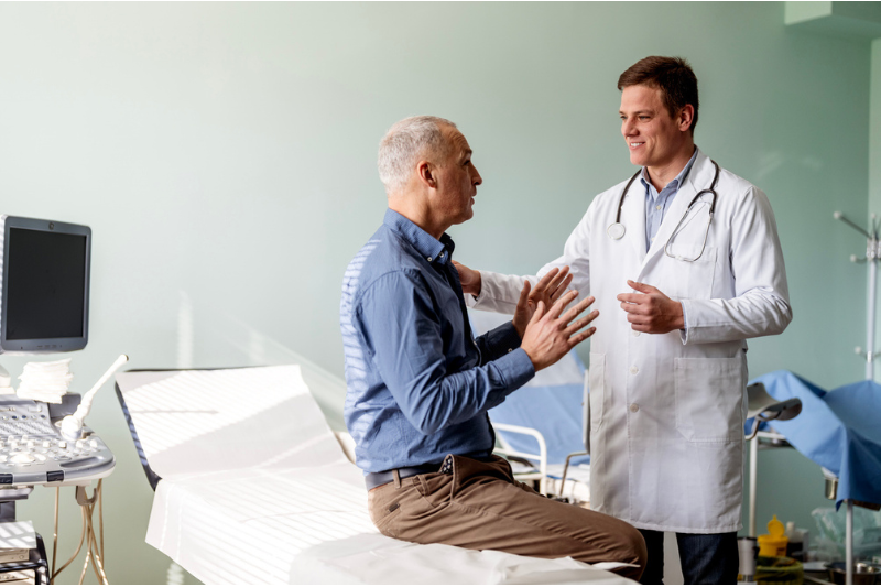 A male doctor stands and listens to his male patient talk.
