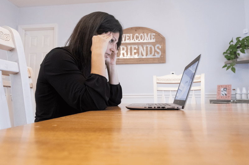 A stressed-out woman holds her hands to her head as she looks at her computer.