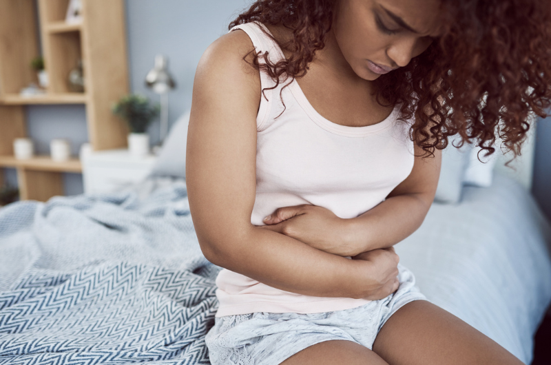 Young woman sits on the edge of her bed and clutches her lower stomach.