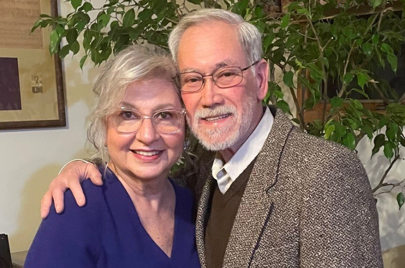 Breast cancer survivor Nora Ambros smiles at the camera while sitting next to her husband.